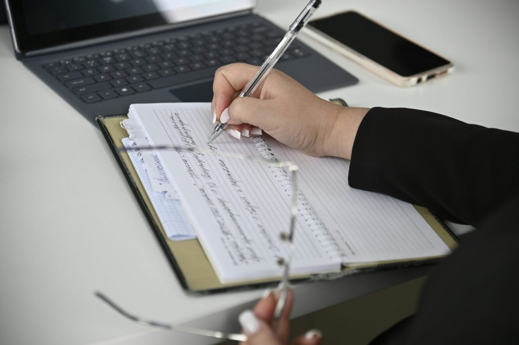 A person writing on a notebook with a laptop in the background
